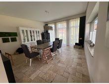 Esszimmer mit großem Holztisch und mehreren gepolsterten Stühlen, daneben ein weißer Vitrinenschrank an der linken Wand. Helle Fliesen auf dem Boden und große Fenster mit transparenten Vorhängen, die viel Tageslicht hereinlassen.