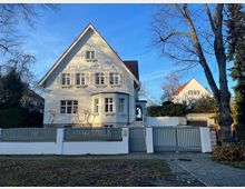 Großes Einfamilienhaus mit heller Fassade und Satteldach, umgeben von einem Zaun und einer kleinen Gartenanlage. Im Vordergrund ein gepflasterter Gehweg, daneben ein Baum ohne Laub; im Hintergrund Bäume und ein weiteres Haus mit roten Dachziegeln.