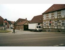 Straßenszene in einer Kleinstadt mit Fachwerkhäusern, einem geparkten weißen Lkw und mehreren Fahrrädern. Im Vordergrund überquert eine Person eine gepflasterte Straße, und "Durchfahrt verboten"-Verkehrsschilder sind sichtbar.