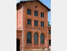Ziegelsteinfassade eines historischen Gebäudes mit großen, rundbogigen Fenstern im Erdgeschoss und kleineren segmentbogigen Fenstern in den oberen Etagen. Der Bau weist dekorative Zierelemente, ein Schrägdach und metallene Fallrohre auf.