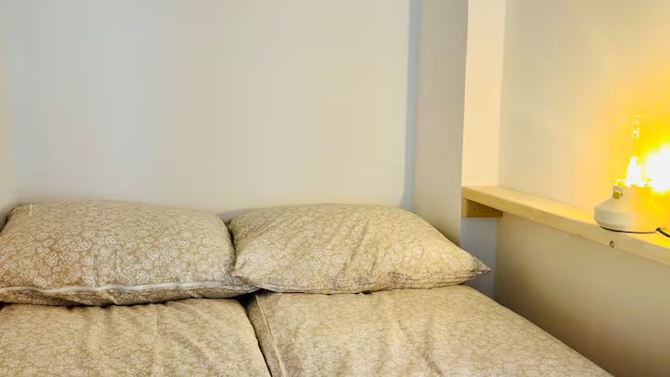 Kleines Schlafzimmer mit zwei zusammengeschobenen Einzelbetten und gemusterten Bettdecken vor einer schlichten weißen Wand. Rechts befindet sich eine gewölbte Wandnische mit Holzablage und einer leuchtenden Tischlampe.