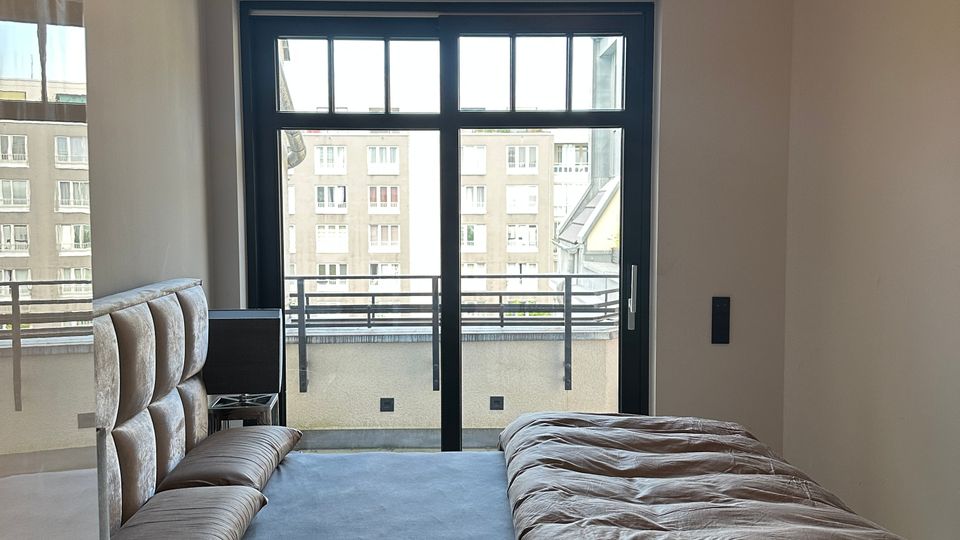 Modernes Schlafzimmer mit Doppelbett und gepolstertem Kopfteil, schlichtem Interieur und ringförmiger Deckenleuchte. Große schwarze Glas-Schiebetür führt auf einen Balkon mit Blick auf umliegende Wohngebäude.