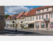 Straßenszene mit einer Reihe von zweistöckigen Wohn- und Geschäftshäusern in heller Putzfassade und roten Ziegeldächern. Im Erdgeschoss befinden sich kleine Geschäfte, darunter ein Blumenladen, und einige Fahrräder stehen auf dem Gehweg; eine ruhige Kopfsteinpflasterstraße führt daran vorbei.