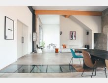 Moderne Wohnung mit offenem Wohnbereich, gekennzeichnet durch helle Wände, sichtbare Holzbalken und eine Glasbodenfläche. Der Raum ist mit minimalistischen Möbeln ausgestattet, darunter ein großer Holztisch, bunte Stühle und ein weißes Sofa, das an einer strukturierten Wand steht.