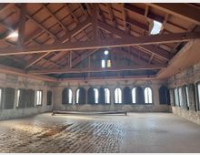 Ein leerer Innenraum mit einem hohen, offenen Holzdachgebälk und Wänden aus rohem Mauerwerk. Der Raum ist von einer Reihe gotischer, bogenförmiger Fenster umgeben, die das Licht hereinlassen, während der Boden mit unebenem Material bedeckt ist.