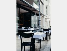 Straßencafé mit schwarzem Mobiliar und weiß eingedeckten Tischen auf einem gepflasterten Gehweg, umgeben von modernen Gebäudefassaden und einer Glasfront. Im Hintergrund sind weitere Tische und ein Sonnenschutz sichtbar.