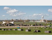 Offene Grünfläche mit einem urbanen Gemeinschaftsgarten aus Hochbeeten und einem Zelt, im Hintergrund das ehemalige Flughafen-Gebäude Tempelhof und ein markanter Radarturm. Der Himmel ist leicht bewölkt, und vereinzelt sind Menschen auf der Fläche zu sehen.