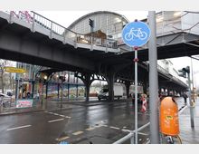 Ansicht einer S-Bahn-Station in Berlin mit erhöhtem Gleis auf einer Stahlkonstruktion. Darunter eine belebte Straße mit Verkehr, Fußgängern und Fahrrädern, sowie ein gelbes Straßenschild mit der Aufschrift "Tiergarten" und bunte Wandmalereien an einem modernen Gebäude.