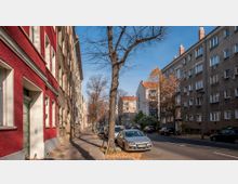 Straßenszene mit Wohnhäusern, die mehrheitlich vier bis fünf Stockwerke hoch sind; linkes Gebäude in kräftigem Rot, andere Fassaden in neutralen Tönen. Geparkte Autos und einzelne Fahrradständer säumen die Straße, während laublose Bäume den Gehweg säumen.