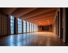 Großer Veranstaltungssaal mit hellem Parkettboden, einer kleinen Bühne am Ende des Raumes und großen Fensterfronten auf der linken Seite, die von gestreiften Vorhängen eingerahmt sind. Die Decke ist orangefarben und wellenförmig, beleuchtet von mehreren hängenden Lampen.