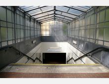 Eingangsbereich zu einer U-Bahn-Station mit Treppen, flankiert von Metall- und Glaswänden im Oberlichtstil. Die Wände sind mit weißen Fliesen verkleidet, und es hängen Hinweisschilder sowie Handläufe an beiden Seiten der Treppen.