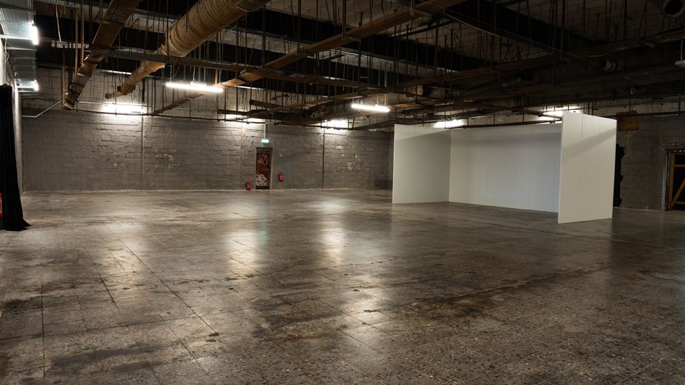 Große, leere Industriehalle mit Betonboden und grauen Steinwänden, beleuchtet von langen Deckenleuchten. An der offenen Decke sind sichtbare Rohre, Kabeltrassen und Lüftungsleitungen, rechts steht eine freistehende weiße Trennwand.