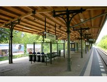 Bahnhof mit überdachtem Bahnsteig im historischen Stil, erkennbar an schmiedeeisernen Säulen und einer Holzkonstruktion. Im Hintergrund Bäume sowie Werbeplakate, der Bahnsteigname "Nikolassee" ist über Sitzmöglichkeiten angebracht.