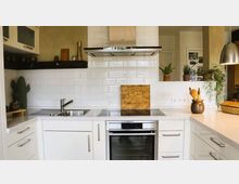 Moderne Küche mit weißen Unterschränken und Arbeitsflächen, einem Herd mit Glaskeramikkochfeld und wandmontierter Edelstahl-Dunstabzugshaube. Die Rückwand ist mit weißen Metrofliesen verkleidet, dekoriert mit Pflanzen und Küchenutensilien.