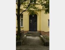 Gelbe Gebäudevorderseite mit einer dunkelgrauen Tür in einem bogenförmigen Eingangsbereich, flankiert von Fenstern. Der Eingangsbereich ist durch eine kurze Steintreppe und einen schmalen gepflasterten Weg mit Bäumen und Pflanzbeeten eingefasst.