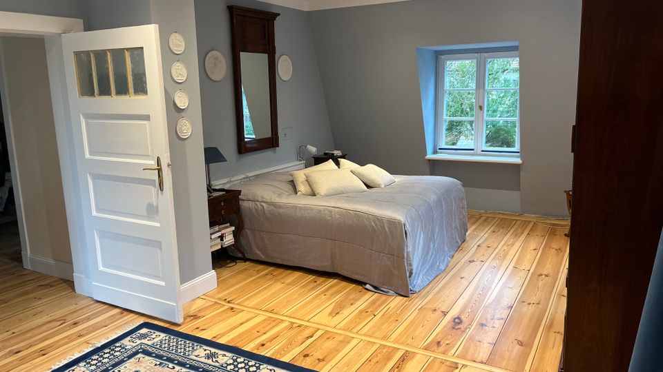 Schlafzimmer mit hellem Holzdielenboden, grauen Wänden und einem Doppelbett mit grauer Tagesdecke; daneben stehen ein Nachttisch und ein hoher Spiegel. Ein weißes Sprossenfenster mit Blick ins Grüne sowie eine offene weiße Tür und dunkle Holzschränke rahmen den Raum.
