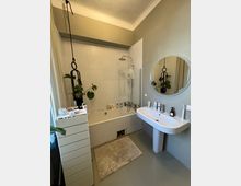 Badezimmer mit einer Badewanne, die mit weißen Mosaikfliesen verkleidet ist, einer daran befestigten Duscharmatur und einer durchsichtigen Trennwand. Rechts ein wandmontiertes Waschbecken mit verschiedenen Pflegeprodukten und einem runden Spiegel darüber; links ein schmaler, weißer Schrank und dekorative Grünpflanzen.