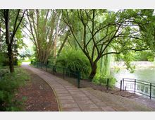 Ein gepflasterter Weg in einem Park führt an Bäumen und Sträuchern vorbei, begleitet von einem grünen Metallzaun. Rechts des Weges liegt ein Teich mit herabhängenden Weidenästen und einer Geländerabsperrung.