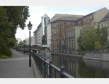 Blick auf einen urbanen Kanal mit angrenzenden Backstein- und modernen Gebäuden. Links verläuft ein Gehweg mit Laternen und Bäumen, während rechts Gebäude und eine Uferwand mit Graffiti zu sehen sind.