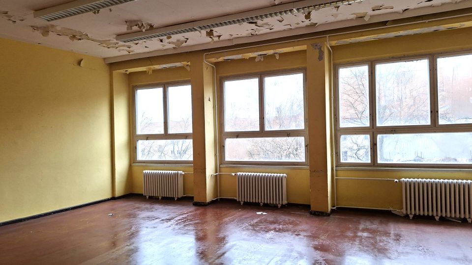 Leerer, renovierungsbedürftiger Innenraum mit gelb gestrichenen Wänden, großen Fensterreihen und Heizkörpern darunter. An der Decke hängen Leuchtstofflampen, und die Farbe blättert großflächig ab; der Boden ist glatt und dunkel.