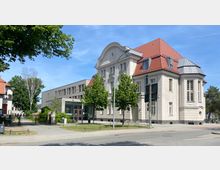 Eckgebäude mit heller historischer Fassade und rotem Satteldach, kombiniert mit einem modernen Anbau aus hellem Beton. Vor dem Gebäude stehen mehrere Bäume, umgeben von einem kleinen Gehweg und einer Straße im Vordergrund.