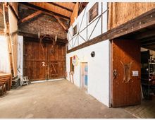 Innenraum einer rustikalen Scheune oder Lagerhalle mit hohen Holzbalken, sichtbaren Dachträgern und teils weiß verputzten Wänden. An den Wänden hängen landwirtschaftliche Geräte und Dekorationen; rechts führt eine offene Holztür in einen Nebenbereich.