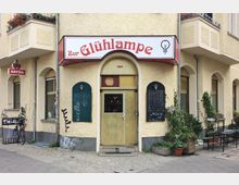 Straßenansicht einer Bar mit dem Namen „Zur Glühlampe“, erkennbar an einem roten Schild über dem Eingang. Die Fassade ist cremefarben und hat Rundbogenfenster, zwei schwarze Tafeln mit Zeichnungen und Texten hängen an der Wand, und im Außenbereich stehen Tische und Pflanzen in Töpfen.