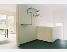 Minimalistisch gestalteter Innenraum mit hellen Wänden, dunklem Bodenbelag und einer schlichten Garderobenleiste an der Wand. Im Hintergrund ist eine Glastür mit Blick ins Grüne zu sehen.