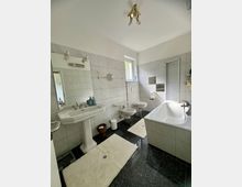 Helles, gefliestes Badezimmer mit großem Waschbecken und Spiegel, Bidet und Toilette sowie einer Badewanne mit Duschabtrennung. Dunkler Steinboden, weiße Badvorleger und ein kleines Fenster sorgen für Kontrast und Tageslicht.