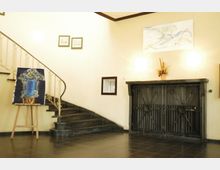 Helle Eingangshalle mit geschwungener Treppe und kunstvoll verziertem Metallgeländer. An der Wand hängen Gemälde und Zeichnungen, während auf einem Holzständer ein farbiges Bild steht; daneben ein dekorativer, dunkler Metallschrank mit floralen Mustern.