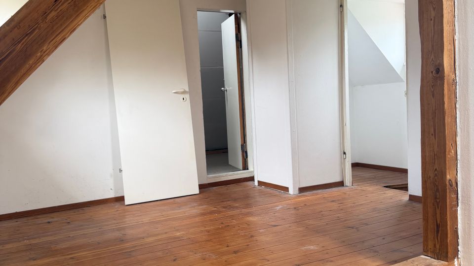 Leerer Dachgeschossraum mit schrägen Decken, sichtbaren Holzbalken und breiten Holzdielen. Weiße Wände mit mehreren offenen Türen führen in angrenzende kleine Räume.