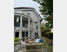 Ein modernes Wohnhaus mit hellem Putz, rotem Satteldach und einem Balkon mit Glasgeländer. Im Vordergrund befindet sich eine möblierte Terrasse mit Stühlen, Pflanzen und Sonnenschirm, umgeben von einem Garten mit Bäumen und Sträuchern.