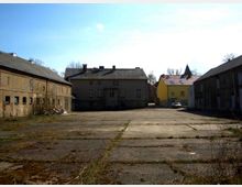 Ein leerer, gepflasterter Hof mit sichtbaren Spuren von Verwitterung, umgeben von alten, teils verfallenen Gebäuden aus Backstein und verputztem Mauerwerk. Im Hintergrund ist ein gelbes Gebäude sowie eine Kirchturmspitze sichtbar.