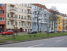 Straßenszene mit mehrstöckigen Wohnhäusern in verschiedenen Farben (Beige, Blau, Gelb und Rot), die sich entlang einer Straße mit parkenden Autos und Bäumen erstrecken. Im Vordergrund befindet sich eine grüne Mittelinsel mit einem Gehweg und einer Sitzbank.