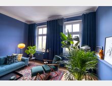 Wohnraum mit blauen Wänden und dunkelblauen Vorhängen, ausgestattet mit einem Sofa, einem grünen Sessel, Pflanzen, einer Stehlampe und einer orientalisch gemusterten Teppichunterlage. Zwei große Fenster lassen Tageslicht herein, und dekorative Elemente wie Bilder und Skulpturen befinden sich auf einer Ablage.