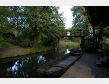 Ein ruhiger Fluss mit einem Holzsteg und einem flachen Boot im Vordergrund, umgeben von dichtem Baumbestand. Eine kleine, hölzerne Fußgängerbrücke überspannt den Fluss und verbindet die Ufer.