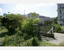 Grüne Parkfläche mit dichten Sträuchern und einer Treppe zu einem unterirdischen Bereich, gekennzeichnet durch ein Schild mit der Aufschrift "HERREN". Im Hintergrund sind Wohngebäude und ein historisches Eckgebäude mit Balkonen zu sehen.