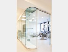 Moderne Arztpraxis mit heller Einrichtung, abgetrennt durch gebogene Glaswände. Im Behandlungsraum stehen ein verstellbarer Untersuchungsstuhl, medizinische Geräte und Schränke vor einem großen Fenster.