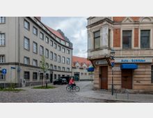 Straßenszene mit gepflasterter Kreuzung, umgeben von zwei Gebäuden: ein größeres, historisches Verwaltungsgebäude links mit grauer Fassade und roten Ziegeldächern, und ein kleineres Geschäftshaus rechts mit orangefarbener Backsteinfassade und blauen Schirmen über Schaufenstern. Im Vordergrund fährt eine Person auf einem Fahrrad.