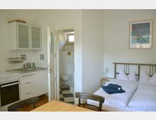 Kleines, helles Apartment mit einer Küchenzeile links, ausgestattet mit Herd, Spüle und Hängeschränken, einem Esstisch im Vordergrund und einem Doppelbett rechts; im Hintergrund eine Tür, die zu einem kleinen Badezimmer mit schwarz-weißem Fliesenboden führt.