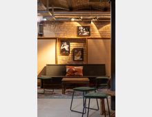 Gemütlicher Innenraum eines Cafés oder einer Lounge mit Ziegelwänden, einer dunkelgrünen Holzbank, einem Holztisch und mehrfach gepolsterten Hockern. An der Wand hängen schwarz-weiße Porträtbilder unter freiliegenden Metallrohren und einer gewölbten Decke.