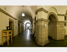Langer, gewölbter Innenkorridor eines historischen Gebäudes mit Steinarkaden und massiven Pfeilern; an den Wänden befinden sich Türen und Beschilderungen, im Hintergrund ist eine Glastür erkennbar. Ein kleiner Holzstehtisch und eine Sitzbank stehen entlang des Weges.