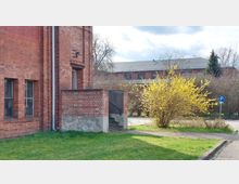 Backsteinbau mit roten Ziegelwänden und schmalen Fenstern, daneben eine kleine Betontreppe mit Geländer. Im Vordergrund ein gelb blühender Strauch, Rasenflächen und Gehwege, im Hintergrund weitere Backsteingebäude und kahle Bäume.