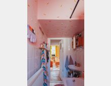 Kleines Badezimmer mit rosa Decke und bunten Konfetti-ähnlichen Dekorationselementen. Im Vordergrund sind Handtücher, ein Waschbecken und eine Badewanne zu sehen, im Hintergrund führt eine Tür in ein helles Wohnzimmer mit gelben Vorhängen und orangefarbenen Möbeln.