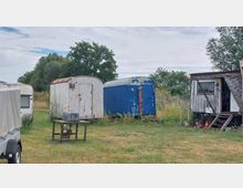 Eine Wiese mit mehreren alten Bauwagen, darunter ein weißer und ein blau gestrichener Wagen, sowie eine wettergegerbte Holzhütte mit Treppe. Im Hintergrund sind Bäume und Sträucher zu sehen.