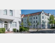 Mehrstöckige Wohnhäuser mit hellen Fassaden und roten Ziegeldächern stehen an einer ruhigen gepflasterten Straßenecke, umgeben von kleinen Bäumen und Sträuchern. Der Bereich wirkt gepflegt und wird von einem klaren, blauen Himmel überdacht.