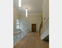 Hell erleuchtete Inneneinrichtung eines Flurbereichs mit hellgrünen Wänden, einem Holzboden und einer hölzernen Tür am Ende. Zu den architektonischen Elementen gehören ein geschwungener Treppenaufgang rechts mit weißen Geländern und ein großes Bogenfenster auf der linken Seite, das Tageslicht hineinlässt.