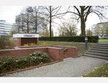 Zugang zu einer öffentlichen Damen-Toilette in einem Park mit Backsteinmauern, Treppen und gepflastertem Weg, umgeben von Bäumen und Sträuchern. Im Hintergrund sind moderne Gebäude und eine grüne Rasenfläche zu sehen.