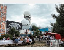 Eine Außenaufnahme eines urbanen Geländes mit einem großen Wandgemälde eines Tigers, mehreren Graffiti-gedeckten Gebäuden und den charakteristischen kugelförmigen Radarkuppeln der ehemaligen Abhörstation auf dem Teufelsberg in Berlin. Im Vordergrund sitzen Menschen an langen Tischen, während weitere Personen vor einem roten Zelt stehen.