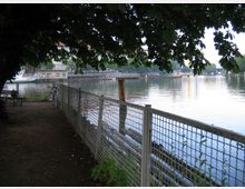 Ein ruhiger Weg unter Bäumen entlang eines Flusses, mit einem Metallzaun, der den Weg vom Wasser trennt. Im Hintergrund sind ein Boot, Gebäude und eine Brücke am gegenüberliegenden Ufer zu sehen.